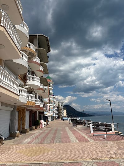 The image captures a scene along the waterfront in Kiparisnoe, Ukraine. A multi-story building with numerous balconies, featuring white ornate railings, lines the left side of the frame. The building’s facade is a mix of tan and brick-like textures, with some balconies showing signs of wear. Shops with white roll-up doors are visible at the ground level.

A paved walkway with a distinct pattern of red and pinkish-yellow diagonal bricks runs parallel to the building and the sea. To the right of the walkway is a wooden fence with a white railing, separating it from the water. A dark body of water, presumably the sea, stretches across the right side of the image, meeting a distant mountainous shoreline. The sky above is a dramatic mix of dark, ominous clouds and patches of bright blue sky with scattered white clouds. A street lamp is visible near the fence. In the distance, a solitary person sits on a bench by the sea. The overall impression is that of a coastal town with a mix of residential and commercial properties, under a changeable sky.
