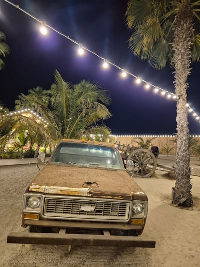 This image depicts an outdoor scene at night in Fujairah, United Arab Emirates. In the foreground, a rusty, old Chevrolet pickup truck is parked on sandy ground. The truck is heavily weathered, with significant rust and damage visible on its exterior, particularly on the hood. The front grille and headlights are intact, and a prominent Chevrolet emblem is visible on the grille.
Behind the truck, to the right, is a large, weathered palm tree. A string of illuminated bulbs is strung overhead, casting a warm light on the scene and extending across the frame. In the background, a rustic wooden cart with large wheels is present. Beyond the cart, there is a low wooden fence and more palm trees. Some of these palm trees are illuminated with lights, suggesting a decorated outdoor venue. The overall environment appears to be sandy and possibly a recreational or dining area. The sky is dark, indicating nighttime. There are no people visible in the image.