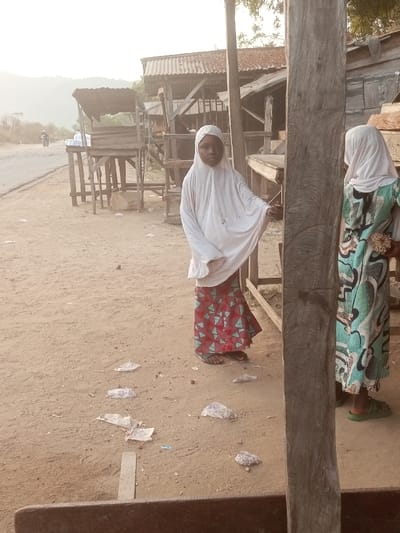 Two young girls wearing hijabs stand outside. One wears a white hijab with a red and green patterned skirt, while the other has a similar white hijab and a teal patterned dress. Trash litters the ground around them. Behind them are wooden structures, possibly market stalls, and a glimpse of a road with a motorcycle. The scene appears to be a rural or developing area.