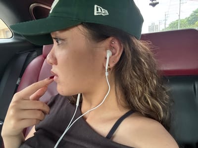 The image shows a young woman sitting in the passenger seat of a car. She is wearing a green baseball cap with a white "NEW ERA" logo, a black shirt, and has long, curly brown hair with blonde highlights. She has her finger on her lip and is looking to the left side of the frame, as if listening to something. Her right ear has three earrings, two gold hoops and one small stud. She is wearing white earphones and has a cord running down her neck and chest. The car seat is burgundy and the car door is visible on the left. The image was taken in the Xinyi District of Taipei City, Taiwan, with the coordinates 22.785 degrees north and 120.297 degrees east.