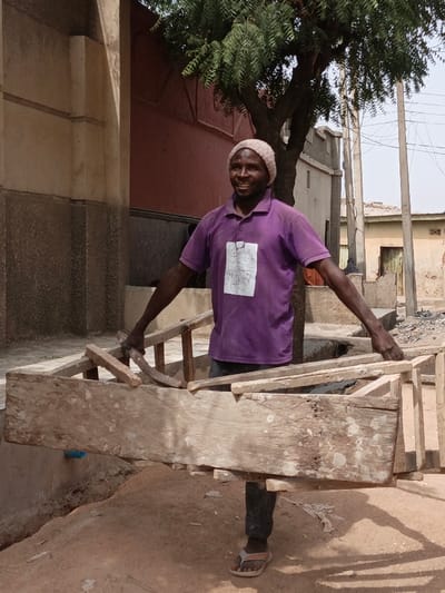 An adult male stands outdoors in a residential area of Zango, Nigeria, centrally positioned and facing the camera with a smiling expression. He wears a light-colored beanie, a short-sleeved purple polo shirt with a faded white rectangular patch on the chest, dark trousers, and light-colored flip-flops. His arms are extended horizontally, supporting a large, rectangular wooden frame constructed from rough, untreated lumber. The ground beneath him is an unpaved, uneven surface of reddish-brown dirt. In the background, to the left, a light-colored building with decorative architectural features is visible. A reddish-brown wall and a large tree with green foliage are present behind the man. To the right, several residential buildings of various construction styles, utility poles, and overhead wires complete the urban environment. The scene is illuminated by bright daylight, casting distinct shadows.