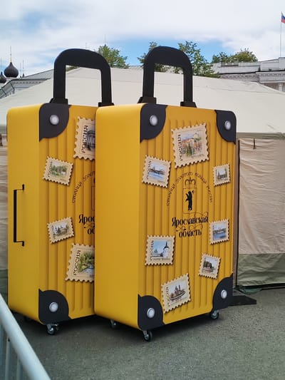 Two oversized, bright yellow suitcases with black handles and wheel bases are displayed outdoors. Each suitcase is adorned with multiple postage stamp-like stickers featuring images of Russian landmarks and text, some in Cyrillic. The suitcases are positioned next to each other, with the right suitcase slightly in front of the left. In the background, white tents and buildings with dark domes are visible under a cloudy sky. The scene appears to be a public event or exhibition in Yaroslavl, Russia, potentially related to tourism in the Golden Ring region, as indicated by the text "Золотого Кольца России" (Golden Ring of Russia) and "Ярославская область" (Yaroslavl Oblast) on the suitcases.