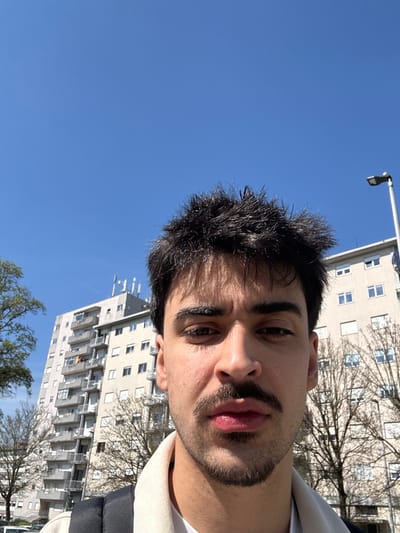 The image depicts a close-up, slightly low-angle portrait of a young adult male outdoors in Guimarães, Portugal, under a clear blue sky. The male subject, centered and facing forward, has dark, spiky hair, dark eyebrows, a mustache, and a short beard. He wears a light-colored collared shirt over a white undershirt, with a dark backpack strap visible over his left shoulder. His expression is neutral.
In the background, to the subject's left, a tall, light-colored multi-story building with numerous balconies and windows is prominent. Clothes are visible drying on at least one balcony, and multiple communication antennas are fixed to the building's roof. To the right of the subject, another similar light-colored building with multiple windows is visible. Several deciduous trees with bare branches are positioned between the foreground and the background buildings. A grey lamppost with a dark fixture extends upwards on the far right side of the frame. The sky is uniformly clear and blue, with a faint white contrail visible in the upper left quadrant. Partially visible at the bottom left are the tops of parked vehicles.