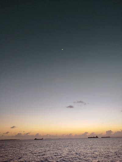 A crescent moon is visible in the dusky sky over the ocean in Gulhi Falhu, Maldives. The sky transitions from a deep gray at the top to a soft yellow and orange hue near the horizon, indicating either dawn or dusk. Several cargo ships are silhouetted on the water, some appearing larger and closer than others. Small clouds are scattered across the sky, some illuminated by the receding or approaching sunlight. The ocean surface is rippled, reflecting the muted colors of the sky. The overall scene conveys a sense of calm and vastness.