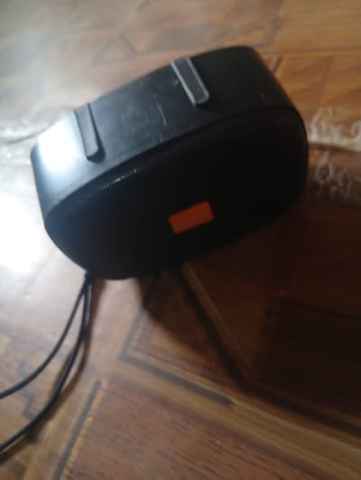 The image shows a black portable speaker with an orange rectangular logo on its surface, resting on a wooden floor with a visible wood grain pattern. Two black cables are connected to the speaker and extend out of the frame. The lighting suggests an indoor setting, possibly a room. There are no people or animals visible in the image. The scene is static and does not depict any action or interaction. The location in the city of Gulak, Nigeria, cannot be determined from the visual information provided in the image.
