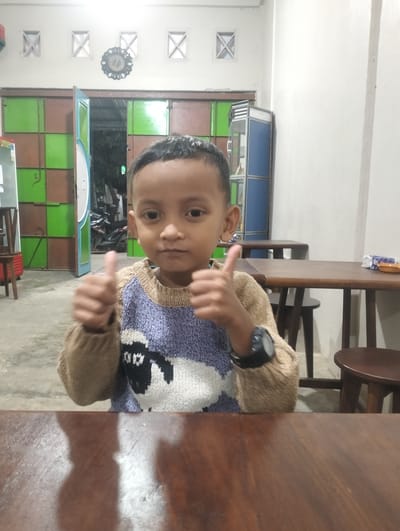 A young child, likely Indonesian given the location context, is the primary subject in this medium shot. They are seated at a wooden table, looking directly at the camera with a neutral expression. The child is wearing a knitted sweater with a pattern of sheep and geometric shapes in purple and brown. A watch is visible on their left wrist. Their thumbs are both raised in a "thumbs up" gesture.
The background suggests an indoor setting, possibly a restaurant or cafe. To the left of the child, there are doors with a checkerboard pattern of green and brown panels. Further back, an open doorway reveals motorcycles and greenery, indicating an outdoor area or courtyard. To the right, more wooden tables and stools are visible, with a light-colored wall and ceiling. A decorative clock with an ornate frame hangs on the wall above the patterned doors. The lighting is artificial, creating shadows and a slightly dim atmosphere, suggesting it is evening or indoors with limited natural light. There is no visible text in the image.