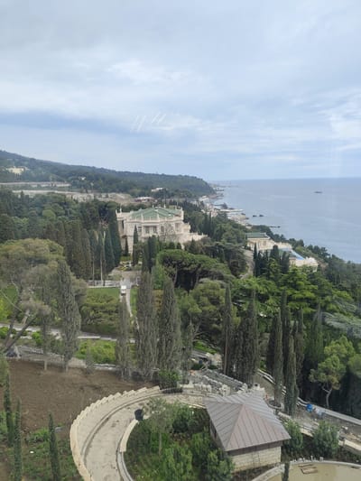 This image depicts a high-angle view of a coastal landscape. The foreground features trees, a stone pathway, and a building with a brown roof under construction. Beyond this, the landscape transitions into dense green foliage interspersed with white and green-roofed structures. In the background is the Black Sea meeting the coast, with a pale sky above. In the distance, a sliver of land is visible on the horizon. The scene seems to capture the Simeiz area in Ukraine, judging by the architectural style and geographical characteristics.