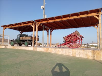 The image depicts a sunny outdoor scene with two vintage vehicles displayed under a wooden-roofed structure. On the left is a dark green truck with wooden accents, its tires resting on a raised platform. To its right is a bright red, wooden cart with large spoked wheels, also positioned on the platform. The structure above them features exposed wooden beams and decorative metal scrollwork along the underside of the roof. The platform is made of large, light-colored stone tiles, contrasting with the green ground in the foreground. In the background, there are some buildings and hills, suggesting a rural or semi-arid location. The sky is a clear, bright blue, indicating fair weather and daytime, likely late afternoon due to the long shadows cast on the ground, including the silhouette of a person holding a camera. The overall impression is one of a historical display or roadside attraction in Albox, Spain. There is no visible text in the image.