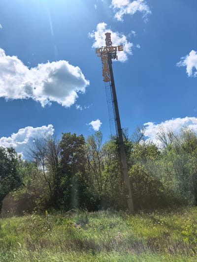 An outdoor, eye-level shot captures a tall, slender concrete pole with a ladder attached, supporting a sign with yellow lettering against a bright blue sky and scattered white clouds. The sign features the words "КАФЕ" (cafe) in large block letters, with a smaller "К" also visible. Trees and green foliage form a dense line behind the pole, and tall, dry grasses and wildflowers grow in the foreground. The image has a slightly hazy quality, possibly due to lens flare. The scene is located in Vozrozhdenie, Russia.