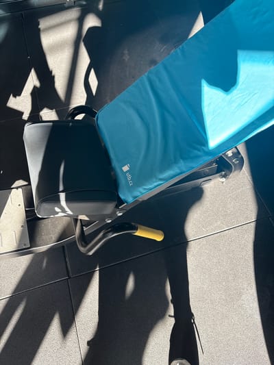 The image shows a piece of exercise equipment, likely a leg extension or curl machine, in a gym or fitness facility. The equipment consists of a padded seat and leg support, a lever with a yellow grip, and a footplate. A light blue towel with "utb.cz" printed on it is draped over the seat. The floor is made of dark gray tiles. Harsh sunlight casts sharp shadows across the floor and the equipment. The location is in a gym facility. The city of Córdoba, Spain, cannot be confirmed by this image.