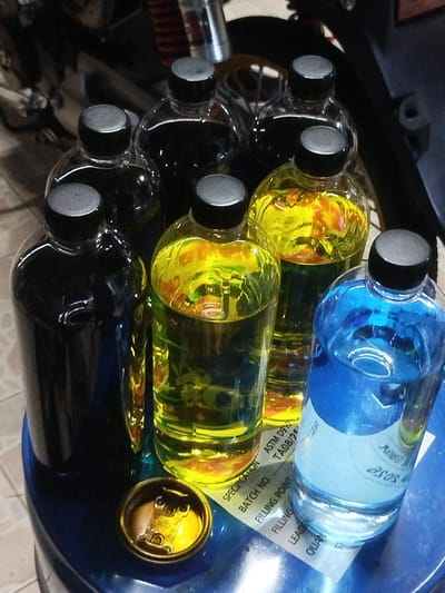 This media file shows a collection of bottles containing various colored liquids, primarily dark, yellow, and blue. The bottles are arranged on a blue surface, possibly a table or stool, with some placed on a piece of paper that has text printed on it. The text visible on the paper includes "SPECIFICATION", "ASTM D910", "BATCH NO", "TA08/2", "FILLING POINT", "FILLING", "LEADE", "QUAN", and "RETEST", as well as the word "asos". The lighting suggests an indoor setting, likely artificial, and the overall impression is that of a product display or storage. There are no people visible in the image. The background is dark and somewhat blurred, hinting at other objects or machinery, possibly related to automotive or industrial use, given the visible text on the paper. The composition focuses on the arrangement and colors of the liquids in the bottles.