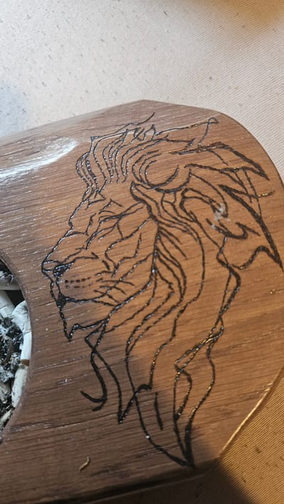 The image shows a close-up of a wooden surface with a pyrographic design of a lion's head. The lion's head is depicted in profile, facing left, with a detailed mane. The pyrography is rendered in black lines. The wood appears to be a medium brown color with a visible grain. To the left of the lion, there is a small pile of what appears to be ash and possibly cigarette butts, indicating a possible ashtray or a surface used for smoking. The background consists of a textured, light-colored fabric, possibly a tablecloth or a backdrop. The scene is photographed from a slightly elevated angle. There are no people visible in the image. The image does not provide any specific information about the city of Kostenets, Bulgaria, or any context related to that location.