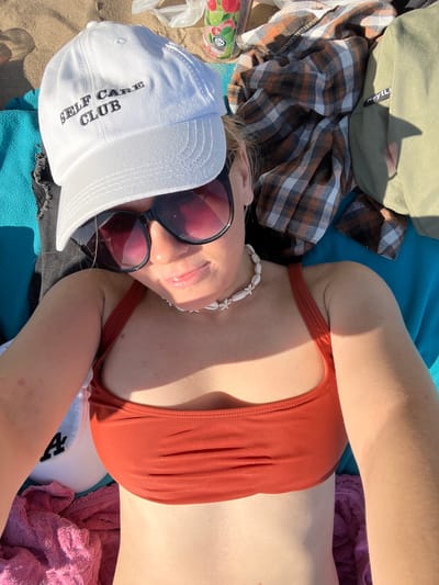 A woman is lying down on a beach, possibly on a towel or blanket. She is wearing a white baseball cap with "SELF CARE CLUB" embroidered on it, sunglasses, and an orange bikini top. She is also wearing a shell necklace. There are other items around her, including a plaid shirt and a green jacket. The beach appears to be a sandy area, and the sun is shining. The image was taken in the city of Ust'ye, Russia.
