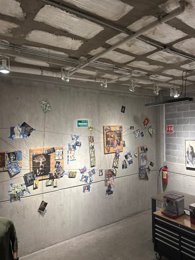 The image depicts an interior space in León, Mexico, characterized by an industrial aesthetic. The ceiling is unfinished concrete with rectangular recessed panels and exposed electrical conduits, from which multiple track lighting fixtures and a linear pendant light are suspended. The left wall is smooth gray concrete, hosting a large, intricate display. This display features numerous small photographs and printed images, secured with blue painter's tape and interconnected by a web of thin dark strings, some attached to three larger wooden panels within the arrangement. A green sign with white text reading "RUTA DE EVACUACION" (Evacuation Route) and an arrow points left above the display. The right wall is constructed from gray concrete blocks and features a red fire extinguisher mounted near the corner. A framed artwork, depicting a monochrome portrait, hangs further along this wall. In the lower right foreground, a black mobile utility cart with multiple drawers and a light-colored wooden top holds a red and silver espresso machine. Part of a dark wooden table extends from the right, behind the utility cart. The lower left corner shows the partial back of a person wearing a dark olive green long-sleeved shirt. The floor is smooth, unpainted concrete.