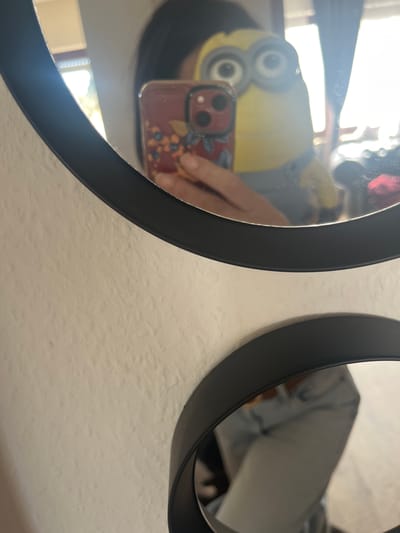 The image shows two circular mirrors with black frames mounted on a white wall. The left mirror reflects a person holding a phone with a red case and floral design, taking a selfie with a Minion plush toy placed on their head. The reflection also shows a window. The right mirror reflects the corner of a white object. The setting appears to be indoors, potentially a domestic environment. The city of Konz, Germany is not discernible from the provided image.