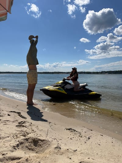 In Hampton Bays, New York, a man stands on a sandy beach, facing away from the camera and holding a hat above his head. He is wearing a green long-sleeved shirt and patterned shorts. To his right, a jet ski is partially in the water. A woman is seated on the jet ski, wearing a white top, shorts, a life vest, and a baseball cap. The water is calm, reflecting the bright blue sky with scattered white clouds. The shoreline is lined with trees in the distance.