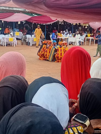 This image depicts an outdoor community event taking place in Ogbogbo, Nigeria. The scene is set under a temporary canopy structure, characterized by draped sections of light pink and dark red fabric overhead. The ground is unpaved, appearing as light brown dirt.
In the immediate foreground, several individuals are visible from behind, facing away from the camera, indicating they are spectators. From left to right, their backs show varied attire: a dark top, a person wearing a pink head covering, another dark top, a person in a dark green top, a white top, a person in a vibrant red head covering, and a person in a white head covering. On the far right, an individual wearing a black head covering is partially visible from the side, with their right hand near their mouth. This person wears a multi-colored patterned sleeve (yellow, black, red, blue) on their left arm, and an Apple Watch with a light-colored band is visible on their left wrist.
In the midground, a group of approximately five to six individuals are crouched or kneeling on the dirt ground. They are dressed in elaborate, matching traditional or ceremonial attire, predominantly yellow and red with distinct striped patterns. This group appears to be performing or engaging in a structured activity. To the right of this group, a man in a blue long-sleeved shirt and green-and-white striped pants stands, observing the activity. Further to the left, a man in a full yellow traditional garment walks across the midground, with his back partially turned to the viewer.
The background reveals an audience area where numerous people are seated at white plastic tables covered with white tablecloths. Small plastic bottles, likely containing water, are visible on these tables. Some tables bear yellow signs with text; partially legible examples include "S.U.O." and "Alum." The attendees are dressed in a mix of traditional and casual clothing. The canopy structure is supported by visible poles, and the overall environment suggests an organized public gathering.
