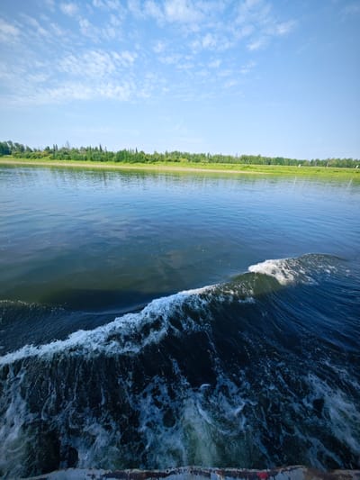 A wide shot captures a body of water, possibly a river or lake, with a distinct wake in the foreground indicating movement. The water is a deep blue-green, reflecting the sky above. A foamy white wave crashes on the right side of the frame, trailing into turbulent water. In the distance, a lush green shoreline is visible, lined with trees and vegetation under a bright blue sky dotted with scattered white clouds. The scene suggests a calm day, possibly during summer, with the perspective implying the photo was taken from a moving vessel. There are no people or animals visible in the image. The specific location is the city of Novonazimovo, Russia.