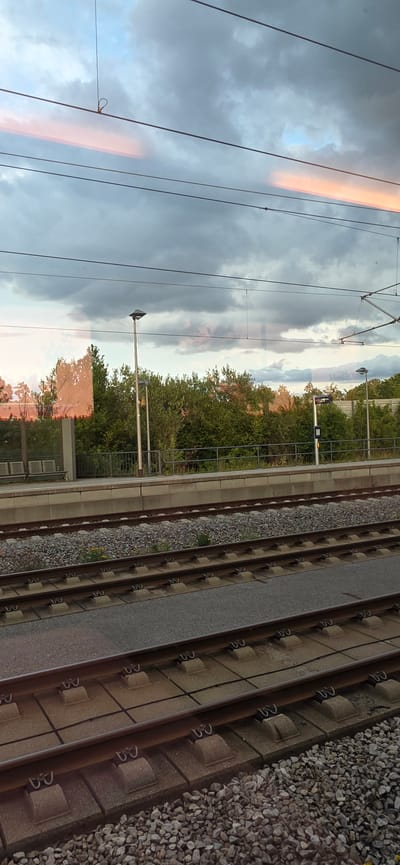 The image shows a train track with ballast and concrete sleepers. The tracks are situated in an outdoor setting, likely a train station or along a railway line. In the background, there are trees and vegetation, suggesting a somewhat rural or suburban environment. Overhead, there are power lines and poles, indicating an electrified railway. The sky is cloudy, with a striking orange or pink hue illuminating parts of the clouds, suggesting either sunrise or sunset. There are also reflections on what appears to be a window or glass surface, showing the sky, trees, and possibly parts of a building. The overall impression is one of a tranquil, yet active, transportation hub at a dynamic time of day. No people are visible in the image. No visible text is present.
