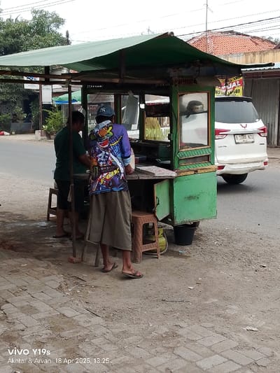 A street food stall with a green canopy stands on the side of a paved road. Two men are standing near the stall. The man in the front, wearing a colorful shirt and brown pants, is looking down at the stall. The man behind him, wearing a green shirt and brown pants, is looking at the side of the stall. A white car is parked behind the stall, facing the street. The stall is covered in a green tarp, and there are several shelves and drawers inside. There is a green metal box attached to the front of the stall. There is a plastic chair on the side of the stall next to the man in the front. The environment is a sunny day with a clear sky and a paved road. The location is Kecamatan Haurgeulis, Indonesia.