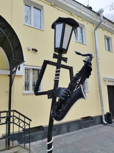 An outdoor, low-angle, eye-level shot captures a black metal sculpture of a lamppost in the shape of a person holding a saxophone. The lamppost has a rectangular lamp at the top, and its body is decorated with white stripes. The saxophone is held by the sculpture's arms, which are also made of black metal. The sculpture is positioned on a sidewalk made of grey paving stones.

In the background, a light yellow building stands. It features several white-framed windows, some with decorative white bars. A black metal railing with circular designs leads up a few steps to an entrance on the left side of the building. A silver downspout runs along the side of the building. Decorative string lights in the shape of musical notes are strung along the building's eaves. The overall environment suggests an urban setting, likely in a public area. The image is taken in Yaroslavl, Russia.