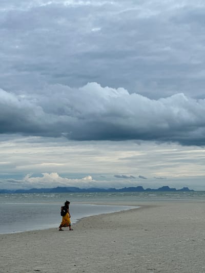 A lone person walks along a sandy beach in Ko Samui District, Thailand. The person, viewed from the side, wears a black shirt and a long, yellow, floral-patterned skirt, and carries a black backpack. They are walking from left to right across the frame, towards the right side of the image. The beach is light-colored sand, meeting a calm body of water on the left. In the distance, a hazy mountain range is visible across the water, under a sky filled with dramatic, dark grey clouds interspersed with lighter patches. The overall atmosphere is overcast and moody.
