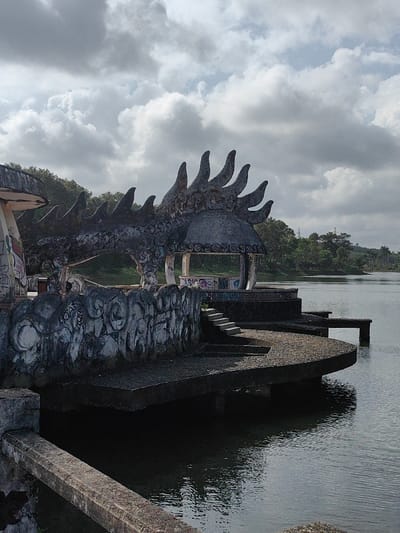 The image depicts a disused waterfront architectural complex located in Kim Long Ward, Vietnam. A large, weathered concrete structure, styled with prominent dragon-like spiny ridges and a head motif, forms an arching canopy over a circular, open-sided pavilion. This primary structure, along with an adjacent ornate concrete retaining wall featuring swirling patterns, exhibits significant degradation, staining, and extensive multi-colored graffiti. Concrete steps descend from the retaining wall to a series of tiered, semi-circular concrete platforms at the water's edge. A narrow, rectilinear concrete pier extends horizontally into the calm, dark-toned body of water from one of these platforms. A second, smaller open-sided pavilion is visible behind the main dragon structure. The background features a dense treeline on the opposite shore of the water body. The sky is predominantly overcast with scattered grey and white clouds. No individuals, animals, or active vehicles are present.