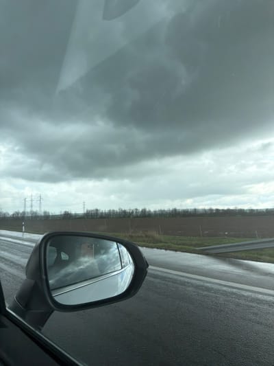 A view from inside a vehicle, likely a car, on a road in what appears to be a rural area under a cloudy sky. A side mirror is visible, reflecting the sky and possibly the vehicle. A strip of trees and fields borders the road. There are power lines visible in the distance. There is no indication that the photo was taken in the city of Vodenichane, Bulgaria. No people or subjects are directly visible. The environment consists of the inside of a vehicle, a wet road, fields, and cloudy sky. The implied action is driving. No interactions are visible.