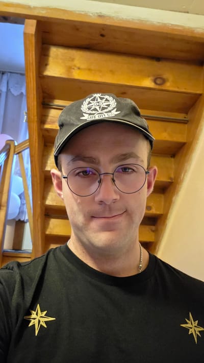 A man with short brown hair and brown eyes, wearing round wire-rimmed glasses, a black baseball cap with a white emblem, and a black t-shirt with a gold star emblem on the left chest, poses for a selfie. He stands in front of a wooden staircase. The scene appears to be indoors.