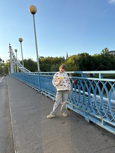 A young person, likely a child or pre-teen, stands on a bridge in Заволжье, Russia, captured in a full-length, daytime portrait. They have light brown hair and are wearing a cream-colored hooded sweatshirt with a red cherry pattern, light grey pants, and patterned clogs. The individual leans casually against a blue metal bridge railing with an intricate, woven-like design. In their hands, they hold a yellow container of popcorn, clearly displaying the text "ВЕСЁЛЫЙ ПОПКОРН" (Cheerful Popcorn).
The scene is set under a clear blue sky with a few faint wisps of clouds, indicative of fine weather. Several white lampposts with spherical light fixtures are visible extending from the bridge structure on the left. Beyond the blue railing, glimpses of dark water are visible, bordered by dense green trees. Distant buildings or spires are partially obscured by the foliage, suggesting a town or city in the background. A small, dark-colored lock is attached to the bridge railing. The bridge's surface is dark grey asphalt or concrete, featuring some cracks and a painted white line. The lighting is bright and even, consistent with late afternoon or early evening.