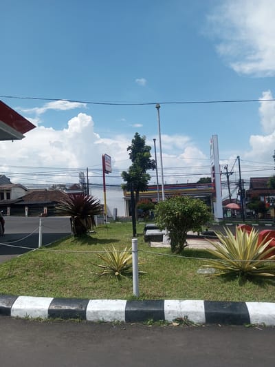 The image shows a street scene on a sunny day. In the foreground is a strip of lawn with various plants, including spiky succulents and small shrubs. The lawn is bordered by a black and white striped curb. A thin rope is strung between white poles, acting as a barrier. In the background, across the street, there are buildings, including a convenience store (Indomaret) and a gas station (Pertamina). Power lines are visible overhead against a blue sky with scattered white clouds. The location is likely in a tropical or subtropical area, based on the vegetation. The specific location cannot be confirmed as Zeyathiri District, Myanmar, without additional information.