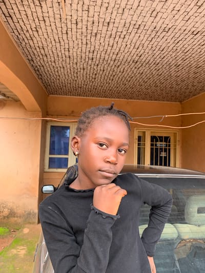 A young Black person with braided hair is pictured from the chest up, in a close-up selfie-style shot. They are wearing a black long-sleeved shirt. Their head is tilted, and they are looking directly at the camera with a neutral expression. A gold earring is visible in their left ear.

The background consists of a brick wall to the left, and a building with orange-colored walls and a window with yellow trim to the right. Some electrical wires are visible hanging from the ceiling. The scene appears to be outdoors, possibly in a residential area, given the brick wall and building structure. The lighting suggests it could be daytime. The overall impression is casual and candid. There is no visible text in the image.
