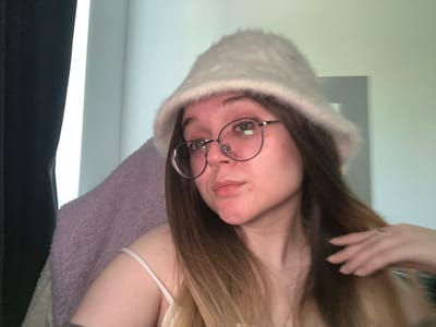 A young woman with light brown hair and glasses wears a white, fuzzy hat. She's also wearing a white tank top and is wrapped in a purple fuzzy blanket. Her right hand is visible near her hair. The background includes a white wall and a dark curtain. The location is not discernible from the image provided, as there is nothing to indicate the location is Pushchino, Russia.