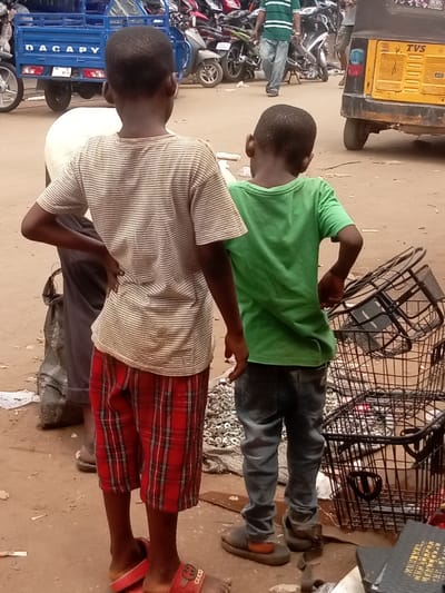 The image depicts an outdoor street scene in Nnewi, Nigeria. In the foreground, two young, dark-skinned males are viewed from their backs, standing on an unpaved, dusty light-brown ground. The boy on the left wears a light-colored, horizontally striped short-sleeved t-shirt, red plaid knee-length shorts, and red slides, one featuring "GUCCI" branding; his hands are placed on his hips. The boy on the right wears a green short-sleeved t-shirt, blue jeans, and dark, chunky slides, with his right hand near his back pocket.
To the immediate left of the left boy, a partial view of an adult in dark trousers is visible. In the mid-ground, a blue cargo tricycle with "DACAPY" written on its side is parked. Behind it, numerous motorcycles and scooters are visible. On the far right, a yellow Keke Napep (tricycle taxi) with "TVS" on its rear is partially seen. Further in the background, an adult male in a green and white striped shirt and blue jeans walks away from the viewer. To the right of the boys, a stack of several black wire baskets is present alongside a small pile of shiny metal components, possibly nuts or washers, resting on a dark surface. Scattered debris, including paper, is present on the ground.