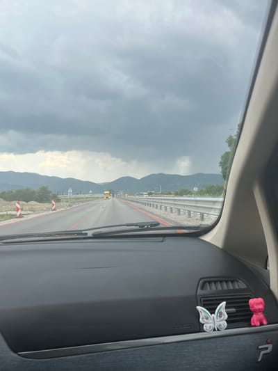 The image, taken from inside a vehicle, shows a highway scene with mountains in the background. The sky is overcast with dark clouds. The road appears wet and has red lane markings. Construction barriers are visible on the left side of the road, and a guardrail runs along the right. A yellow truck and other vehicles are on the highway in the distance. On the car's dashboard, a white butterfly ornament and a pink dog figurine are visible. The location is identified as Skravena, Bulgaria.