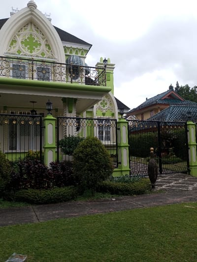 A vibrant green and white house with ornate architectural details, including arched windows and a balcony with wrought iron railings, stands behind a black metal fence. Lush greenery and a well-manicured lawn fill the foreground. In the background, another house with a distinctive tiled roof can be seen under a cloudy sky.