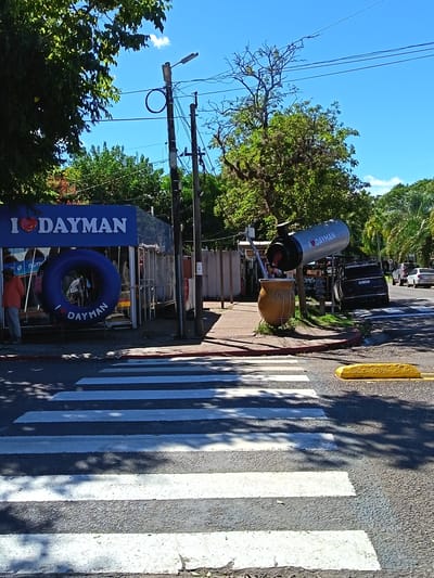 The image shows a street scene in Termas del Daymán, Uruguay. A crosswalk is in the foreground. On the left, there is a shop with a blue sign reading "I [heart] DAYMAN" and a large inflatable inner tube with the same text. A man wearing an orange shirt stands in front of the shop. In the middle of the image, a large yerba mate gourd and bombilla (straw) sculpture is visible, with a cylindrical addition above it with "I [heart] DAYMAN". Beyond this, cars are parked on the street. Utility poles with wires are strung across the sky. Trees provide shade. The sky is blue with few clouds. A yellow parking curb is on the right.