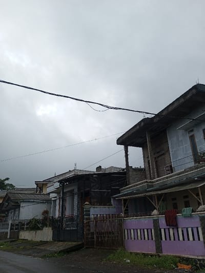 The image depicts a residential street scene in Ambakirano, Madagascar, under an overcast sky. The foreground features a dark, possibly asphalt or dirt, road with patches of green weeds and soil. Along the right side of the road, a low concrete fence with alternating purple and white vertical stripes is visible, adorned with a dark red and a light green cloth hanging over it. To its left, a rustic dark brown wooden gate provides access to a property.
Beyond the fences, a series of houses line the street. The building closest on the right is a two-story structure with unpainted gray walls, featuring a prominent flat-roofed balcony on the upper level supported by thick concrete pillars. Brown-framed windows are visible on both stories. A lower, sloped roof extends from the main structure, supported by wooden poles over what appears to be a ground-level patio area. Utility lines, consisting of multiple dark cables, span horizontally across the upper portion of the image, running above and between the buildings. Further left, additional residential structures are visible, characterized by varied roof types, including corrugated metal and tiles, and predominantly muted color palettes. No individuals are present in the scene, and no overt actions or interactions are depicted. The overall environment suggests a quiet, potentially damp residential area.