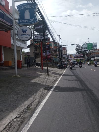 A street scene in Indonesia, with a Lawson convenience store sign prominent on the left. Motorbikes travel on a two-lane road. A man stands on the sidewalk next to parked motorbikes. Overhead power lines tangle across the sky. Advertisements and various signs line the street further down. The sky is overcast.