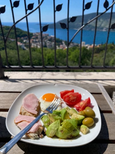 The image shows a breakfast plate on a wooden table outdoors. On the plate are sliced tomatoes, olives, avocado, a fried egg, and ham on bread. A fork rests on the plate. In the background, a wrought iron railing obscures a view of a body of water, trees, and buildings, which appears to be the city of Trebesin, Montenegro.