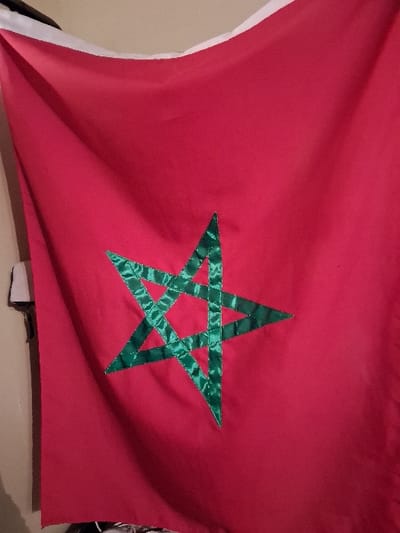 A photograph presents the flag of Morocco. A bright red background serves as the backdrop, contrasting with a large, vivid green five-pointed star at its center. The star appears to be constructed from a glossy, satin-like material, adding a textural element to the design. The flag's left side has a white border. The flag seems slightly wrinkled, possibly indicating its recent use or display.