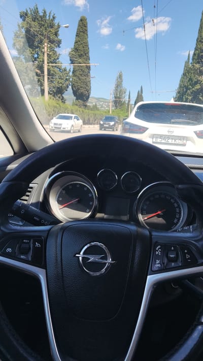 The image is taken from the driver's perspective inside a car, looking forward through the windshield. The steering wheel, dashboard with a tachometer and speedometer, and part of the car's interior are in the foreground. Outside, several cars are visible on a road. The scenery includes tall cypress trees, power lines, and a cloudy blue sky. In the background, there are rolling hills with some buildings, suggesting a town or city. The license plate of the white car in front is visible and reads "E 542 TK 43". Based on the license plate format and common vehicle types, this scene is likely in Russia, not Alushta, Ukraine.