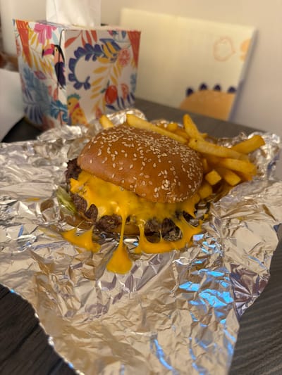 A cheeseburger and fries are placed on a wrinkled piece of aluminum foil, which is situated on a dark, textured surface. The burger is topped with sesame seeds and a generous amount of melted yellow cheese. French fries are visible behind and to the right of the burger. In the background, a tissue box with a colorful tropical pattern featuring toucans and foliage is to the left, and a white surface with a faint, light-colored drawing of a smiling cloud or sun is to the right. The image does not provide any information to determine the location in the city of Leeds, United Kingdom.