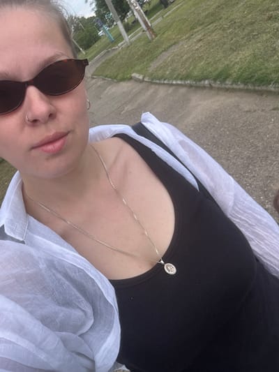 The media file is a close-up, low-angle, outdoor selfie taken by a person wearing sunglasses and a black top with a silver necklace. The person is also wearing a light-colored, sheer garment draped over their shoulders and chest. The background shows a grassy area and a paved path, suggesting an outdoor setting like a park or a sidewalk. The lighting appears to be natural daylight, possibly overcast given the lack of strong shadows. The person's expression is not fully visible due to the sunglasses, but the open mouth and slight tilt of the head could suggest a casual or relaxed mood. There is no visible text in the image. The provided location context is Rozhdestvenskaya, Russia.