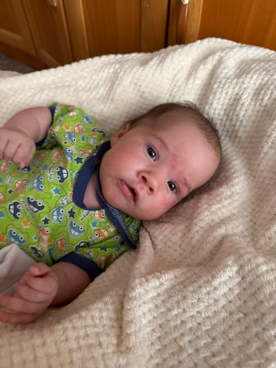 A newborn baby is lying on a fluffy white blanket, looking upwards and slightly to the side. The baby is wearing a green onesie with a print of cars and stars. A wooden cabinet is visible in the background. The scene appears to be indoors, likely in a home environment, with no specific identifying features of the city of Vylgort, Russia.