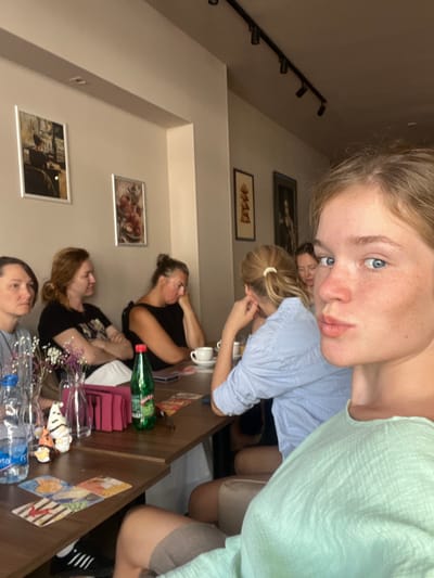 In Donji Pobori, Montenegro, a group of at least six individuals is seated around a wooden table, seemingly in a cafe or restaurant. The perspective is a selfie taken by a young woman in the foreground, who is looking towards the camera with a slightly puckered expression. She is wearing a light green textured top.

Behind her, to her left, another young woman with blonde hair tied in a ponytail is wearing a blue and white striped shirt. Further to the left, a woman with dark hair is looking to the side, her arms crossed. Next to her, a woman with red hair also has her arms crossed and is looking away. Two more women are visible in the background, one with dark hair and the other with blonde hair, both appearing to be engaged in conversation or contemplation.

On the table, there are several objects including bottles of water, a green bottle of what appears to be soda, a white teacup and saucer, and some small decorative gnome figurines. Several cards, possibly tarot cards, are laid out on the table in front of some of the individuals. Pictures hang on the wall in the background, adding to the ambiance of the establishment. The lighting suggests it is daytime.