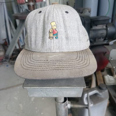A light gray baseball cap with a darker tan brim rests inverted on a flat, L-shaped metallic surface, secured to a vertical metal pole by a visible bolt. The cap features an embroidered depiction of the cartoon character Bart Simpson, wearing a red shirt, blue shorts, and light blue shoes, holding a skateboard. Two dark eyelets are present on the upper front panels of the cap. The background, out of focus, reveals an industrial or workshop environment located in Gimpo-si, South Korea, characterized by various machinery, including a dark electric motor, a reddish-brown cylindrical object, and a gray plastic jerrycan. No individuals are visible.