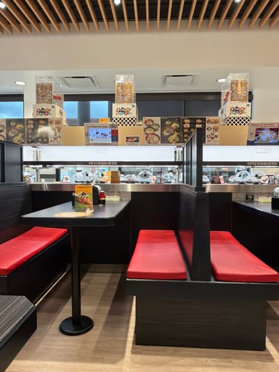 The image shows the interior of a Kura Sushi restaurant in Beaverton, Oregon, USA. The restaurant features a conveyor belt sushi system with individual booths and tables. The booths have red seating and dark wooden tables. On the tables are menus, small condiment bottles, and a yellow flyer for "Kura Reserve". Above the conveyor belt, there are displays of merchandise, including toy prizes and animated advertisements. The overall ambiance is modern and clean, with wooden slat accents on the ceiling. There are no people visible in the image.