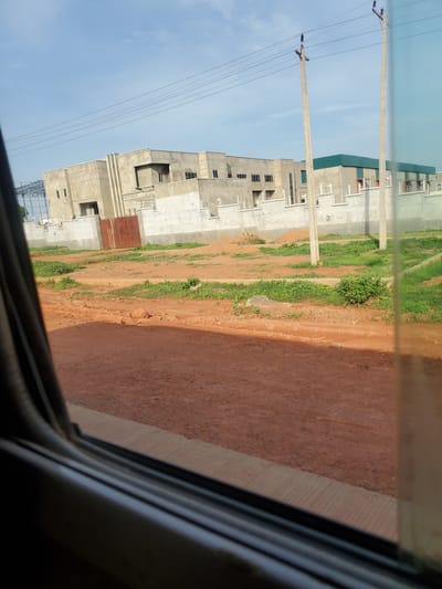 This is a medium-distance, eye-level shot of an ongoing construction project on a sunny day in Kakau, Nigeria. The scene is viewed from inside a vehicle, as evidenced by the dark window frame in the foreground. A partially completed building complex, constructed with concrete, is visible in the background. The building features a light-colored facade with a dark green metal roof on a section of it. A tall, rusty metal gate is situated in front of one of the building's entrances. Several utility poles with electrical wires are also present. The immediate surroundings of the building are cleared land with some patches of green grass. The foreground is dominated by a reddish-brown dirt road. The sky is blue with scattered white clouds.