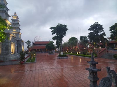 The image shows a wet, brick-paved plaza in Hong Gai Ward, Vietnam. In the background, there is a Ferris wheel. To the left, a multi-tiered stone structure is illuminated with golden lights. In the center of the plaza, there is a manicured tree. Other greenery, including shrubs and trees, are distributed around the plaza. The sky is overcast, and the light is muted. In the foreground, there is a stone sculpture of a pagoda-like structure and a bird.